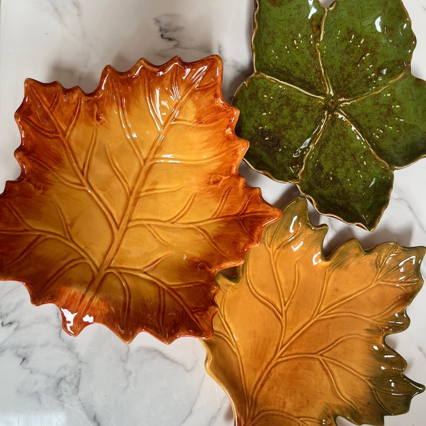 Three ceramic leaf-shaped serving dishes with glossy glazes, displayed on a white marble surface. One dish is orange-brown, one is green, and one is golden yellow.