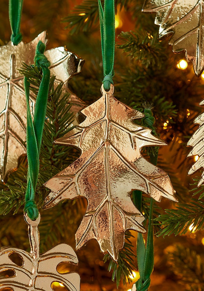 A set of metallic gold leaf-shaped Christmas tree ornaments hanging from a green Christmas tree with warm yellow string lights.