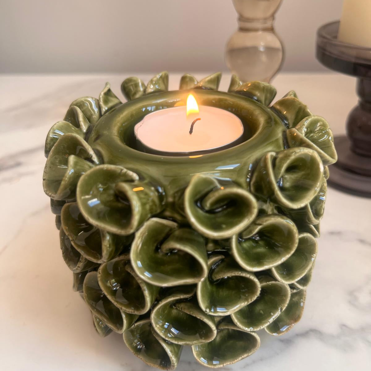 A green glazed ceramic candle holder with a ruffled, lotus-like exterior, holding a lit tealight candle on a white marble surface.