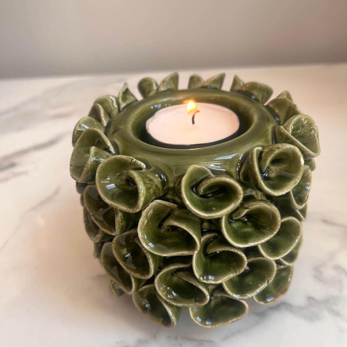 An olive green ceramic candle holder with a textured, scalloped shell-like design, holding a lit white tea light candle on a white marble surface.