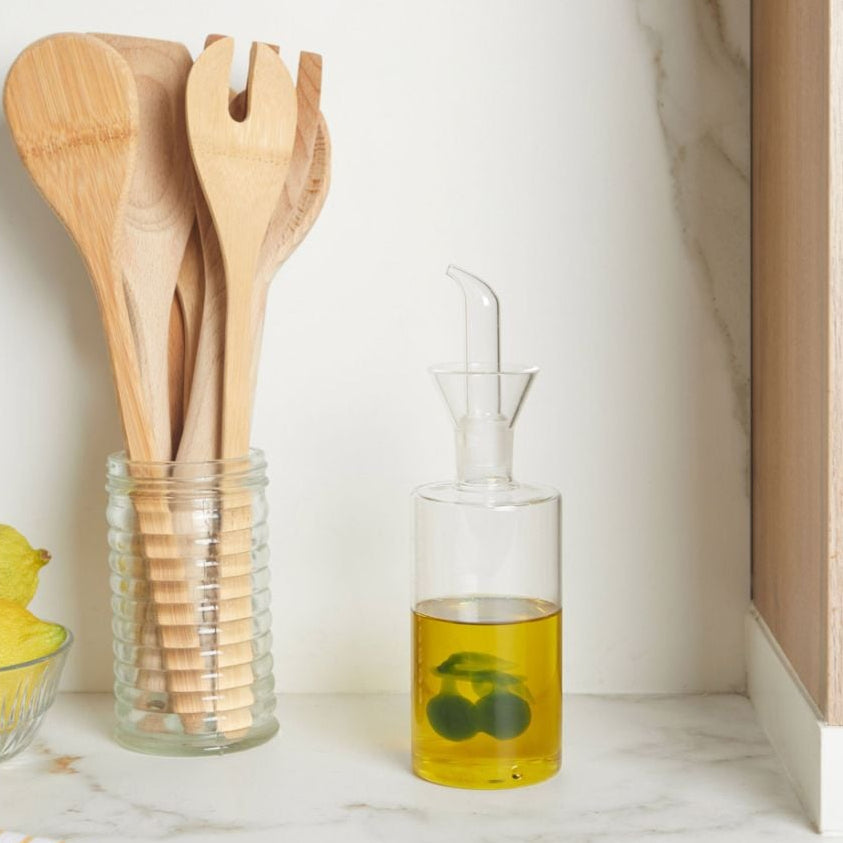 Clear glass container with olive oil and lemons next to a jar of wooden utensils on a marble surface.