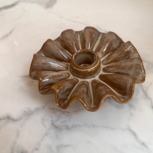 A brown and beige glazed ceramic flower-shaped dish or candle holder with a scalloped rim and a central hole, resting on a white marble surface.