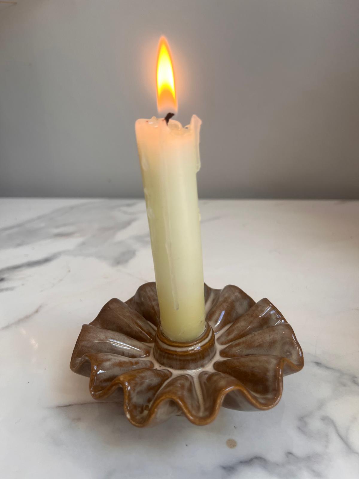 A lit cream-colored pillar candle standing in a brown scalloped ceramic holder on a white marble surface against a gray background.