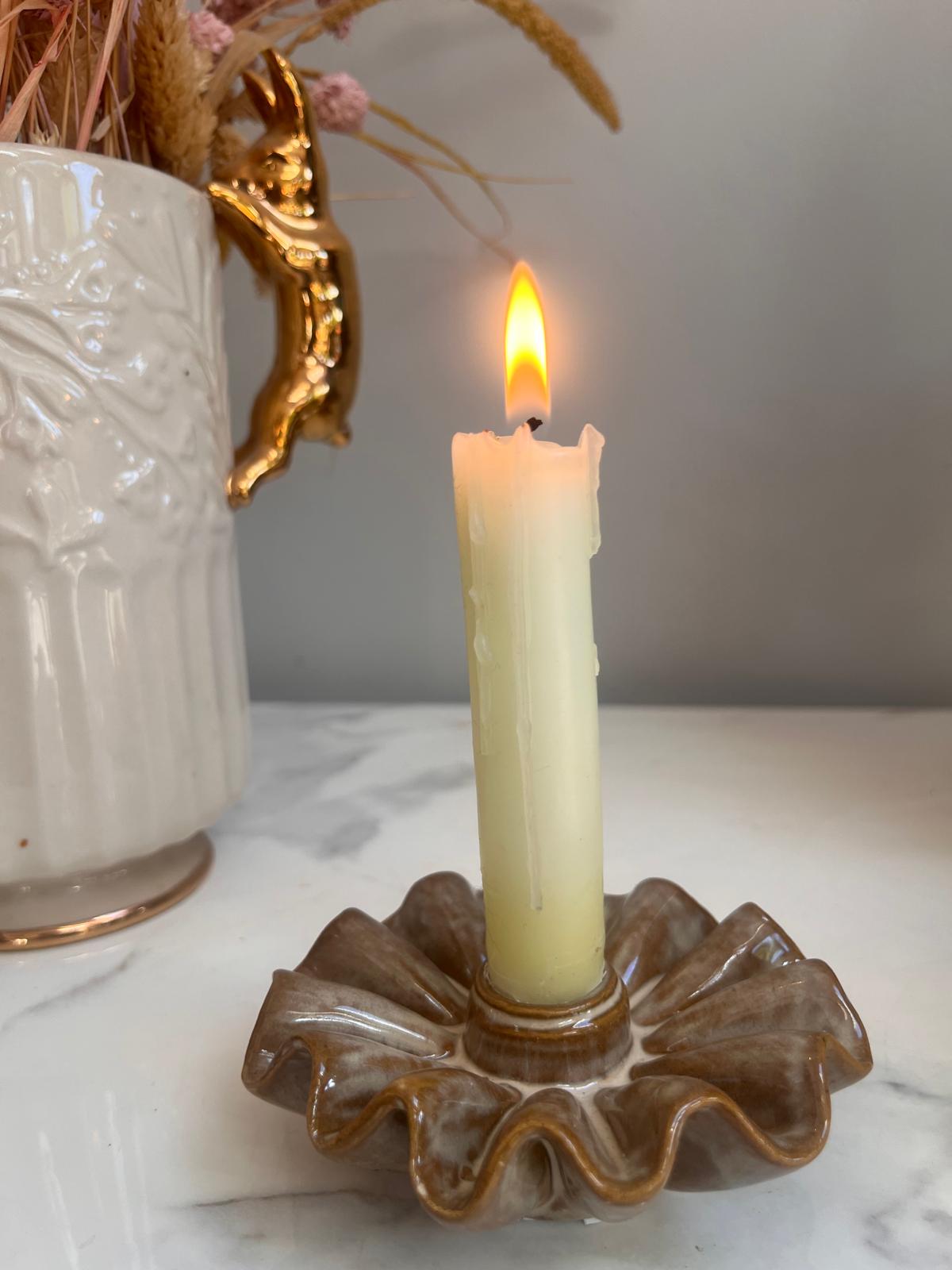 A lit cream pillar candle standing in a brown ruffled ceramic holder on a white marble surface, with a white vase and dried flowers blurred in the background.