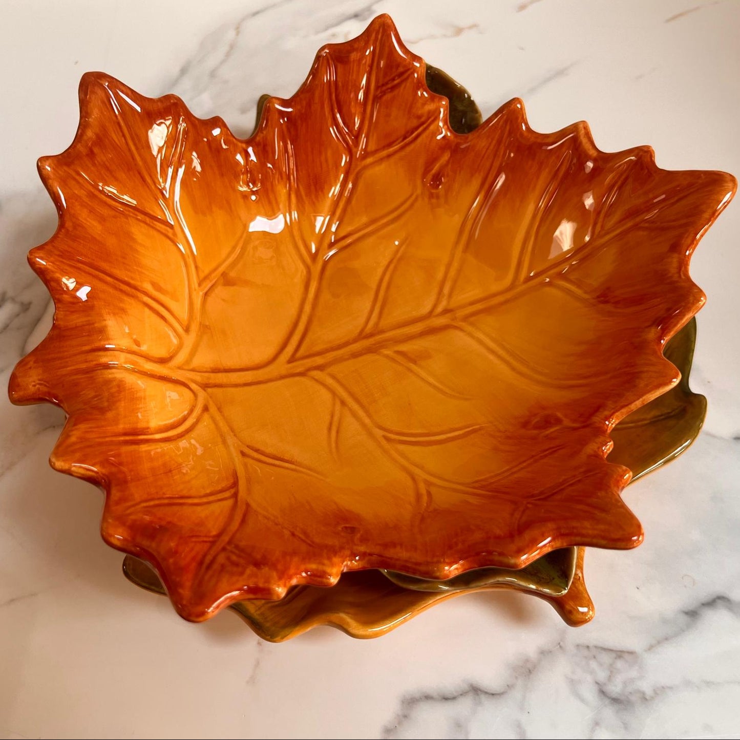 A glossy ceramic maple leaf-shaped bowl in warm orange and amber tones, stacked on another leaf-shaped dish on a white marble surface.
